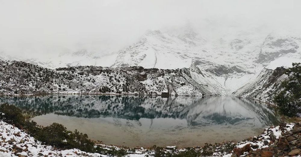A serene mountain lake (Science) surrounded by snow-covered hills and trees (AI), reflecting the snowy landscape and overcast sky (reflections of Science with AI). The lake is clear and calm, with rocky and snowy terrain in the foreground and mist-shrouded mountains rising in the background. Near Cusco, Perú.