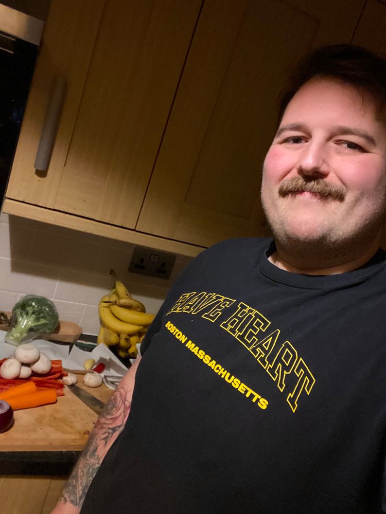 A selfie of me in my kitchen in front of a chopping board stacked with vegetables. I have stubble, a moustache and fluffy hair pushed to one side. 
 I'm wearing a black tshirt with HAVE HEART in a yellow outline varsity style font arched across my chest and BOSTON, MASSACHUSETTS in bold, solid yellow underneath
