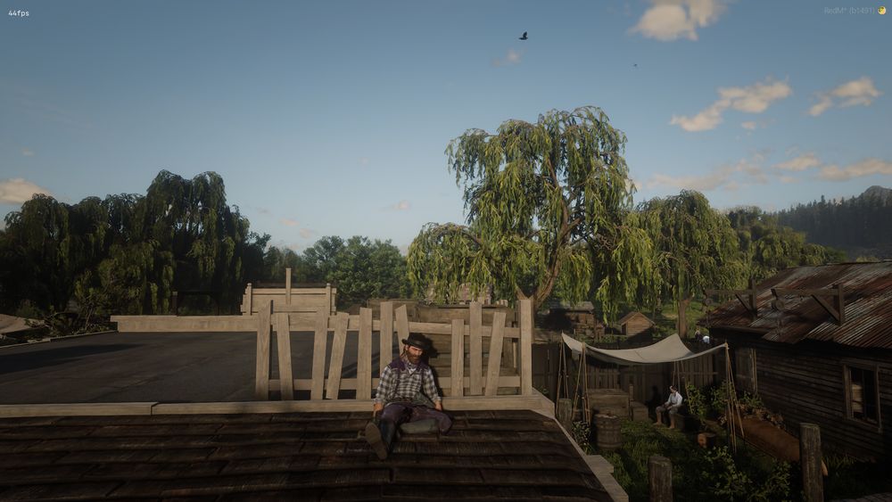 A man in a checkered shirt and dark pants sits slumped on the edge of a rooftop, visibly exhausted. His posture suggests he is regaining strength. Behind him stands a partially constructed wooden structure. The setting is a small rural town surrounded by lush willow trees, under a clear blue sky with scattered clouds. Below, people are gathered near a canvas canopy set up in a grassy yard between wooden buildings.