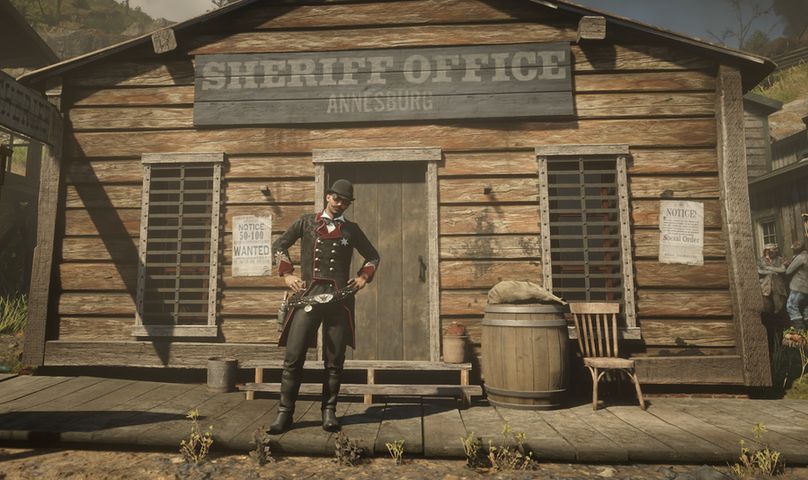 A man stands outside of a sheriff's office on the porch. He's wearing dark clothing with hints of red. He has a moustache, goggles, and what appears to be a bowler styled hat. He stands with his hands on his hips, the sun reflecting off of the Sheriff Badge that is pinned to his chest.