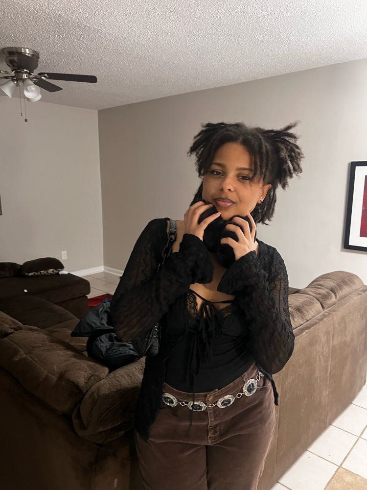 me posing with a black top and brown jeans in front of a brown couch holding ear muffs :)