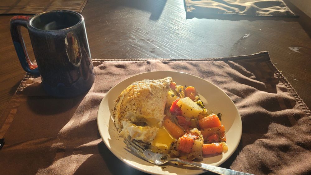 Mug of apple cider next to a a plate of eggs, yolks broken, with roast carrots, potatoes, peppers, and carrots.