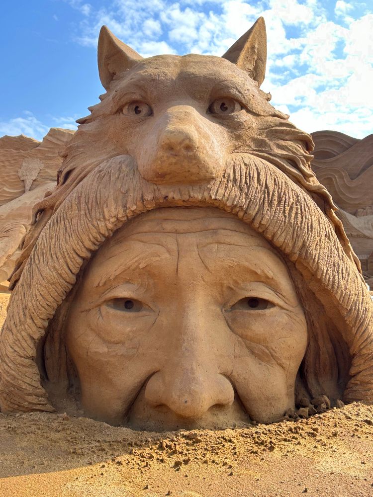 Sandskulptur: Das Gesicht einer alten Frau, bis zur Nase im Sand verschwunden, ernster Blick in die Kamera, über ihrem Kopf ein Wolfsgesicht, darüber blauer Himmel bei Sonnenschein, einzelne weiße Wolken 