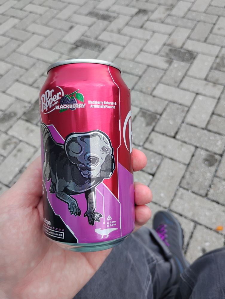 dr pepper special blackberry flavor with purple branding and a cartoon dinosaur (aquilops) on it for a jurassic world rebirth promotion. holding it in my hand over my legs and shoes (they have purple laces) and the grey brick pavement, still a little wet from the rain.