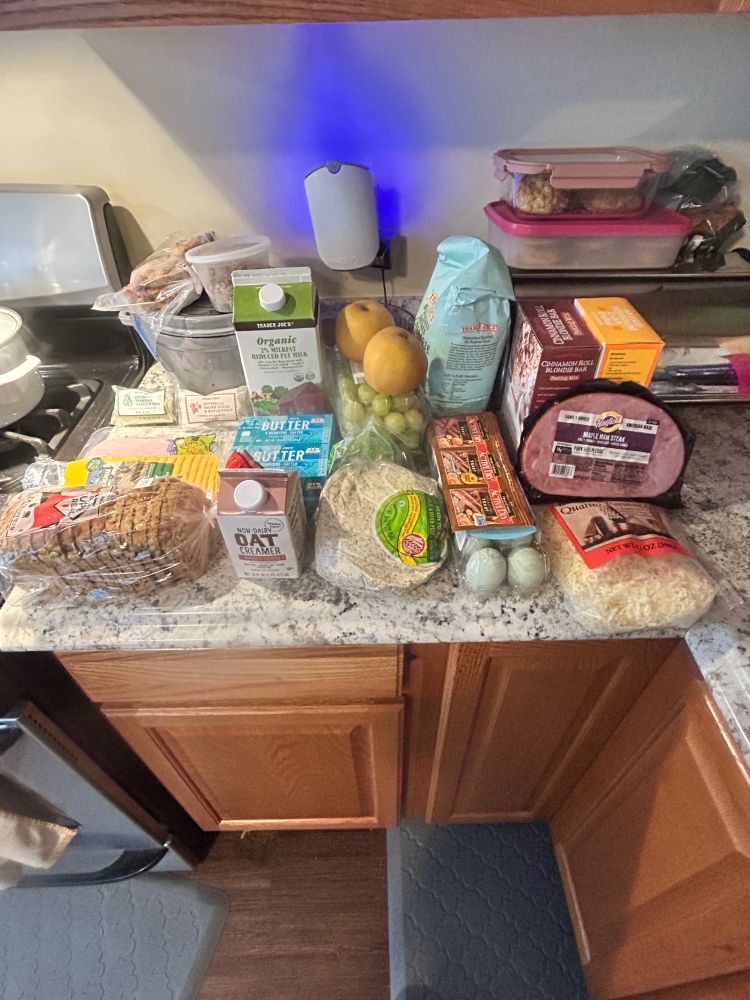 Groceries stacked up on a countertop. There is ham, cheese and bread. Various dairy products like eggs and milk. And fruit like grapes and Asian pears. 