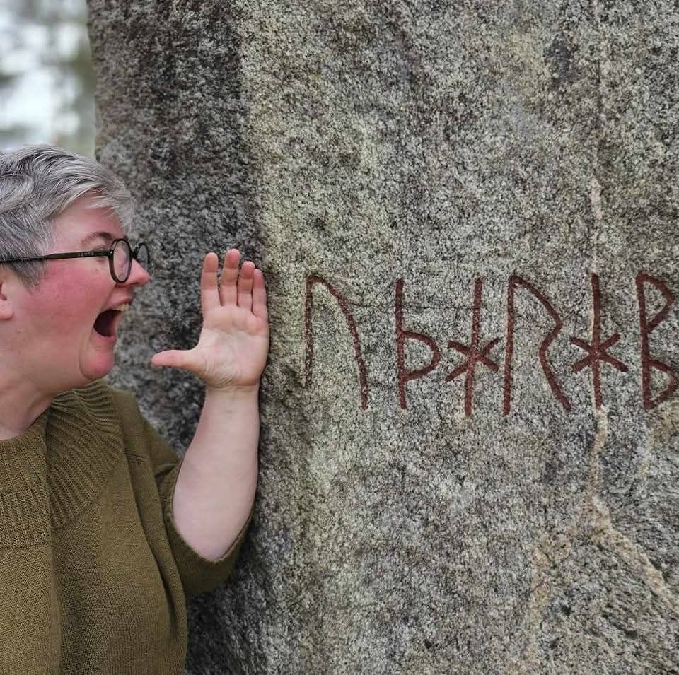 A photo of Maja, a round woman with grey hair and glasses, standing next to a rune stone. She is pretending to yell the runic inscription, which is UÞARABASPA "I prophecy destruction".