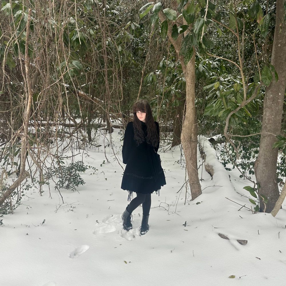 A white woman with long, black hair is wearing a black velvet dress and standing in the snow. The woods behind her are making her look disproportionately small, like a fairy.