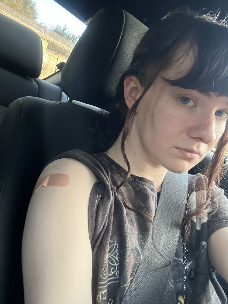 A white woman with black hair and bangs sitting in a car with an unamused expression. Her sleeve is rolled up to reveal an improperly-placed band-aid on her arm, implying she got a shot.