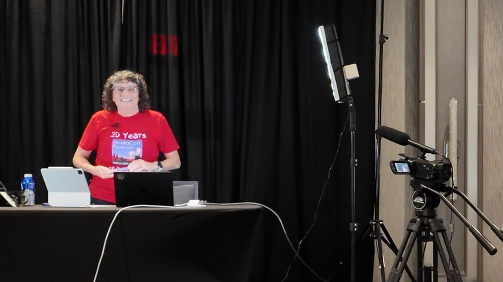 Photo of Allison about to give her presentation on the stage at Macstock 2025. She is smiling broadly and wearing a red shirt that says “20 Years of Podcasting” with the NosillaCast logo in the middle. She is standing behind a desk with a Mac and iPad sitting open on top and a video camera and lighting to her right. 