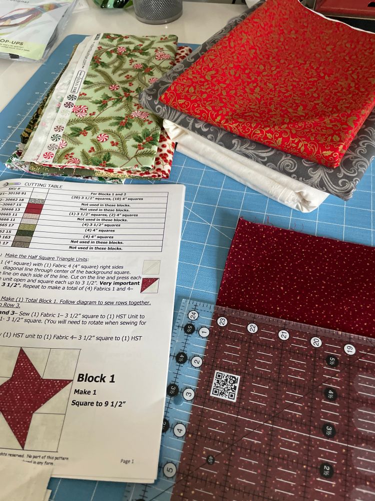 Several cuts of Christmas themed quilt fabrics surrounding a Christmas tree themed quilt pattern on a blue cutting table