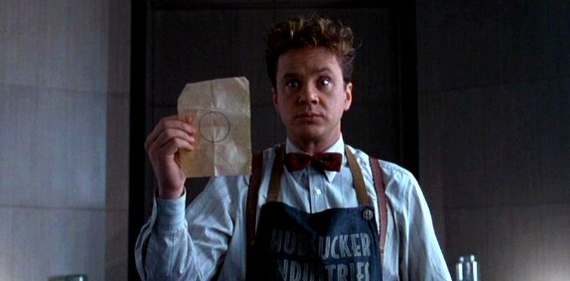 Screenshot from The Hudsucker Proxy. Tim Robbins wears a Hudsucker Industries apron and holds up a ragged piece of paper with a circle drawn on it. 