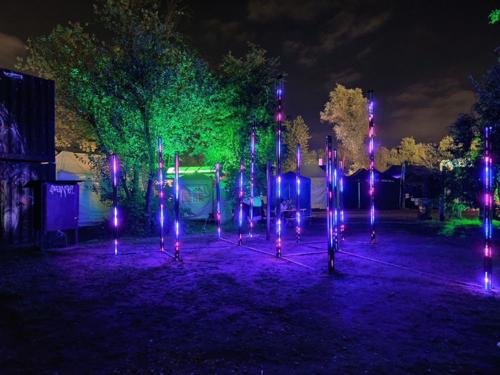 Ein Kunstwerk aus blau-lila leuchtenden Säulen. Dahinter beleuchtete Bäume.