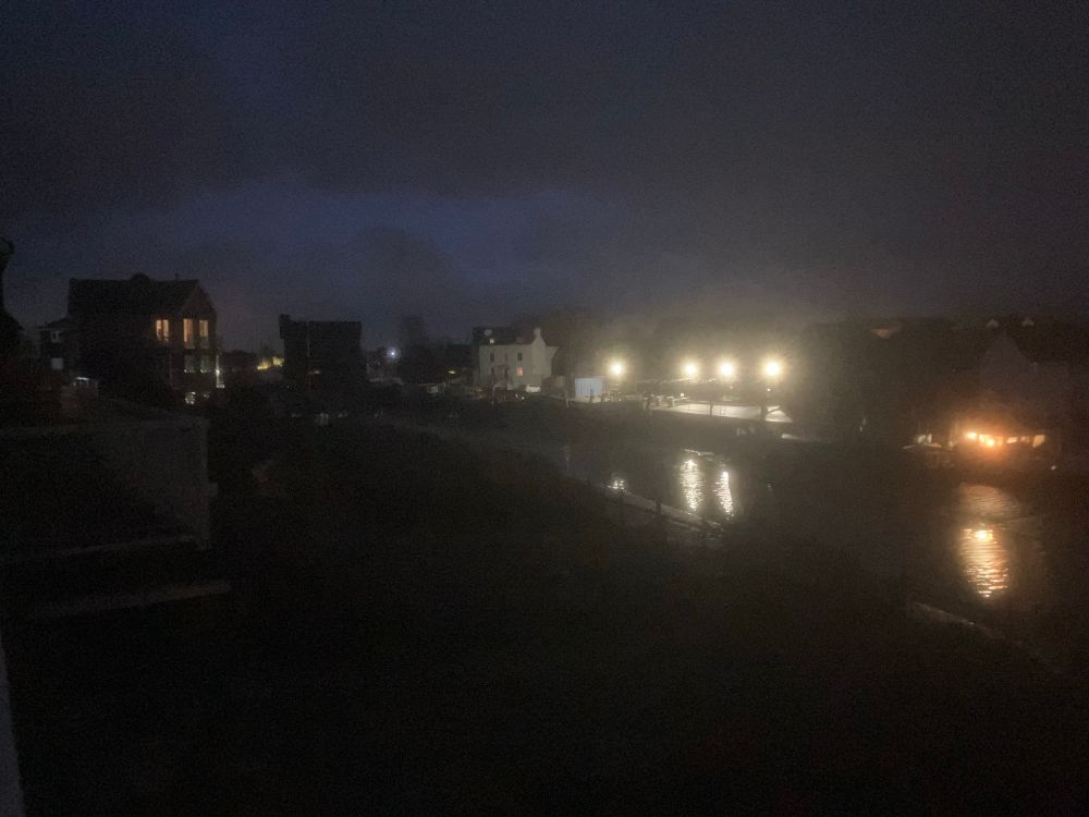 A glimmer of light in the sky to the east - electric lights still bright in front of the buildings across the creek 