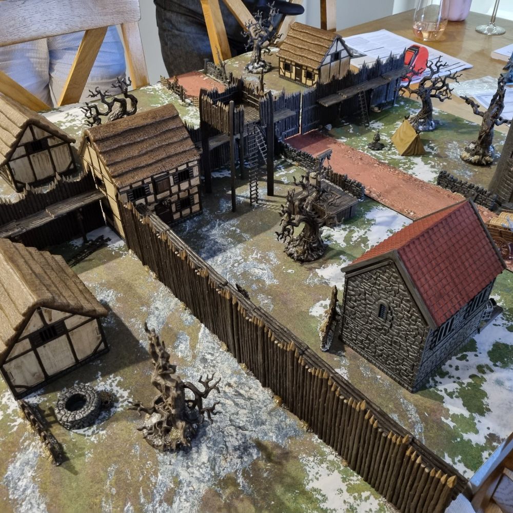 Model village set up for wargaming with barricades, palisades, dead tree and gallows.