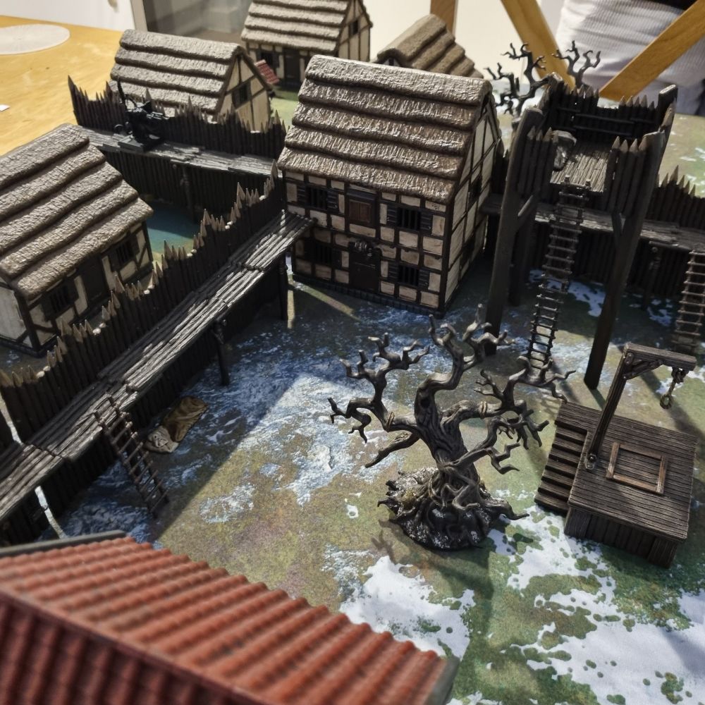 Model village set up for wargaming with barricades, palisades, dead tree and gallows.