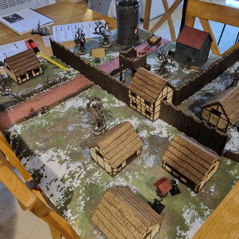 Model village set up for wargaming with barricades, palisades, dead tree and gallows.