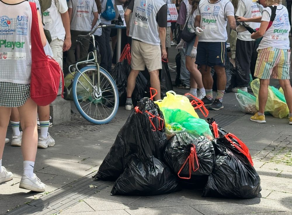 Ein Haufen Müllsäcke liegt auf dem Boden, ringsherum sind Personen in Spogomi-Leibchen zu sehen