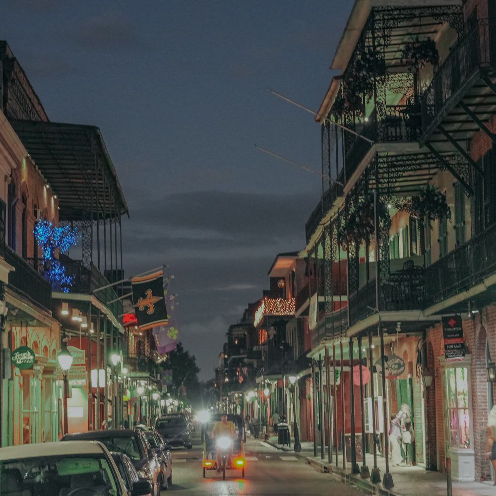 A pedal cab is center frame riding towards you at evening on bourbon street. A New Orleans Saints flag hangs off the balcony of a building on the left. 