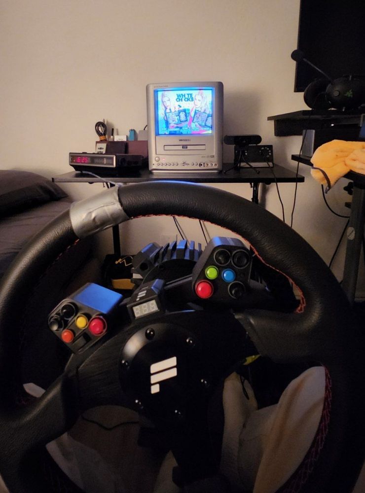 A first person perspective of a dork sitting in a racing simulator seat with his steering when in front of him and an itty bitty crt tv in the background on a desk displaying white chicks on dvd