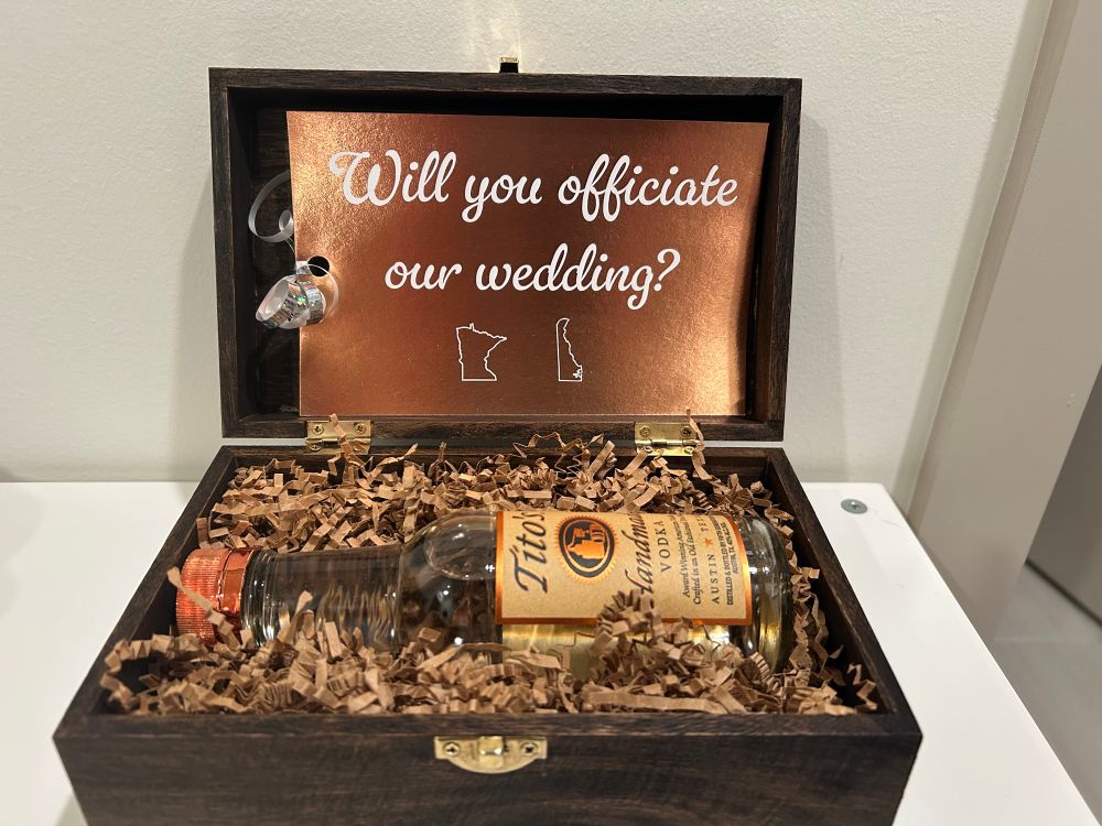 A dark wood box that contains a bottle of Tito’s vodka. A bronze card reads “Will you officiate our wedding?” in a cursive font and the outline of the states of Minnesota and Delaware below it.