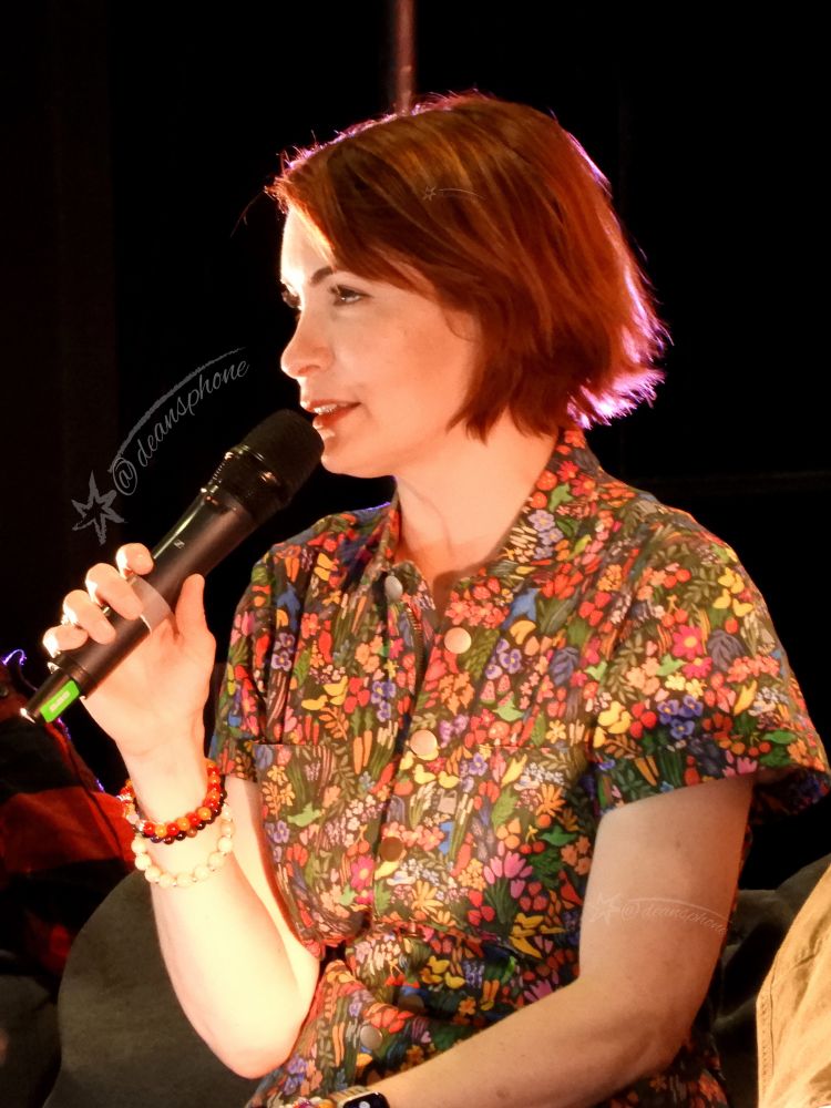 A red haired woman in a colorful jumpsuit. She’s talking with a microphone. 