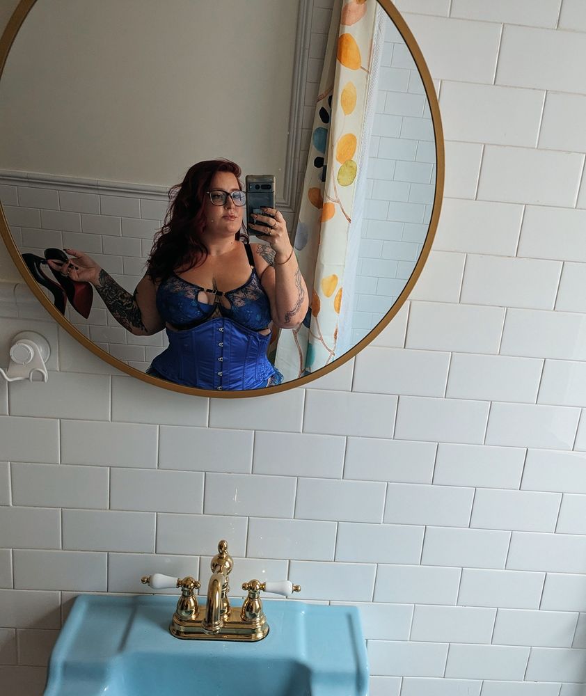 Ava takes a mirror selfie in a round mirror. She is wearing blue lingerie and a blue corset holding red bottoms with one hand.