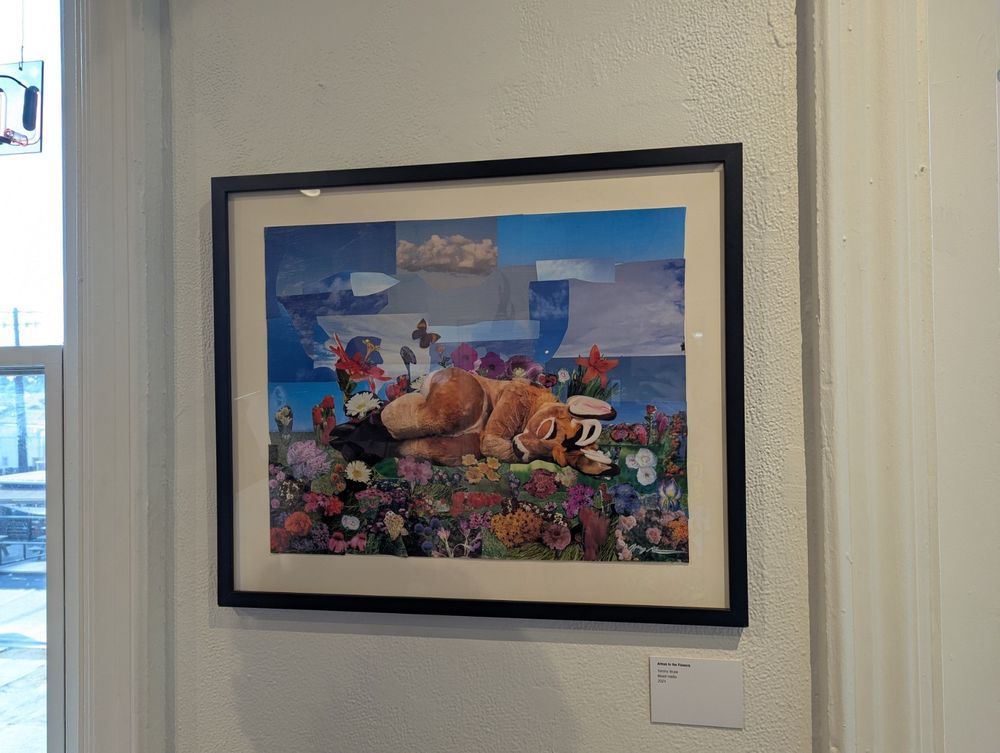 a photograph of a mixed media piece from the Room Party art show. the artwork is titled "Atmus in the Flowers" and shows a photo collage of a fursuit lying down in the flowers.