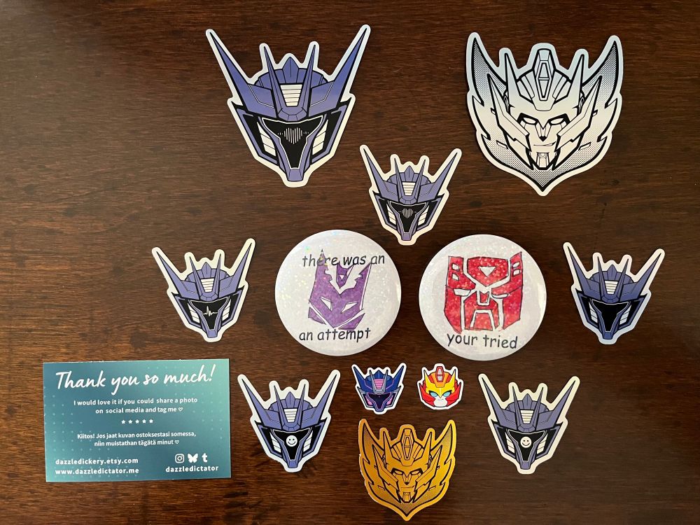 Photo of an assortment of stickers: TFP Soundwave heads with different designs on the visors (heart, soundwave, smiley face), some Rodimus stars, some mini SW and Rodimus heads, and two funny pins drawn in a childlike style with intentional grammar/spelling errors. the Decepticon pin says "there was an an attempt" and the Autobot pin says "your tried."