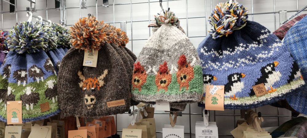 Wollmützen mit Motiv nebeinander. Von links: Wiese mit schwarzen Schafen, einzelne Highlandcow, mehrere Highlandcows, Tiere die Sarah Kutter für süße Pinguine hält 