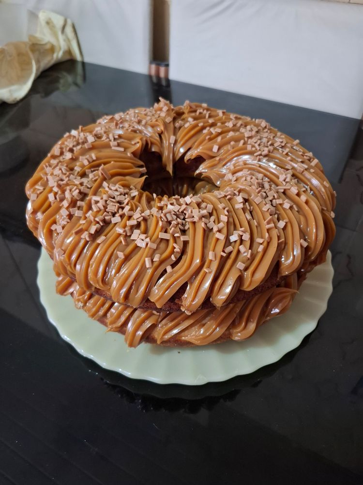 imagem de um bolo todo decorado com doce de leite em buco de confeitar, coberto com granulado de caramelo. ele tem um buraco no meio, e está num prato bege. ao redor tem a mesa de vidro preto e pode-se ver algumas cadeiras de cor creme