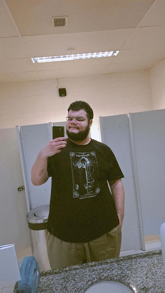 foto minha no espelho (homem branco, gordo, de barba e cabelos pretos, óculos redondo), estou usando uma camiseta preta com uma arte do Nosferatu, um vampiro careca, de orelhas e dentes pontudos, a arte está inteira num tom meio ocre. estou usando uma bermuda bem larga, quase da mesma cor ocre e a mão direita está no bolso dela, enquanto a esquerda segura o celular. atrás de mim estão algumas cabines do banheiro