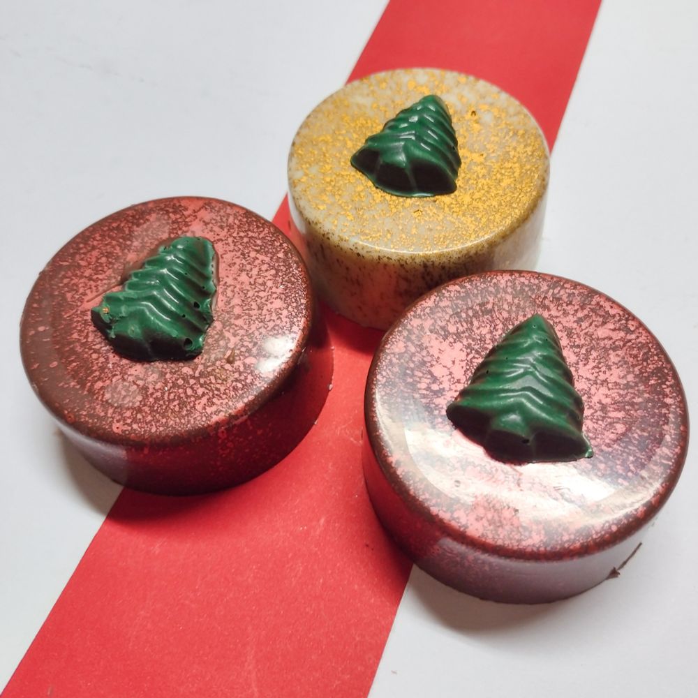 imagem de pães de mel decorados com pó metalizado comestivel, de chocolate branco com decoração dourada e de chocolate ao leite com decoração vermelha. todos eles tem um pinheiro de chocolate tingido de verde em cima

o fundo também é branco e vermelho