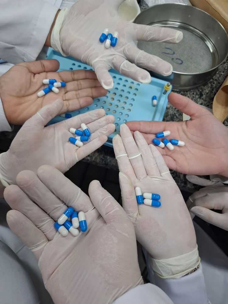 foto com 6 mãos com algumas cápsulas azuis e brancas. as 3 mãos mais próximas do canto inferior da foto e a última do canto superior estão de luva branca, as duas do meio estão sem luva. ao fundo, estão a encapsuladora e a peneira, que tem um nome específico mas esqueci qual é
