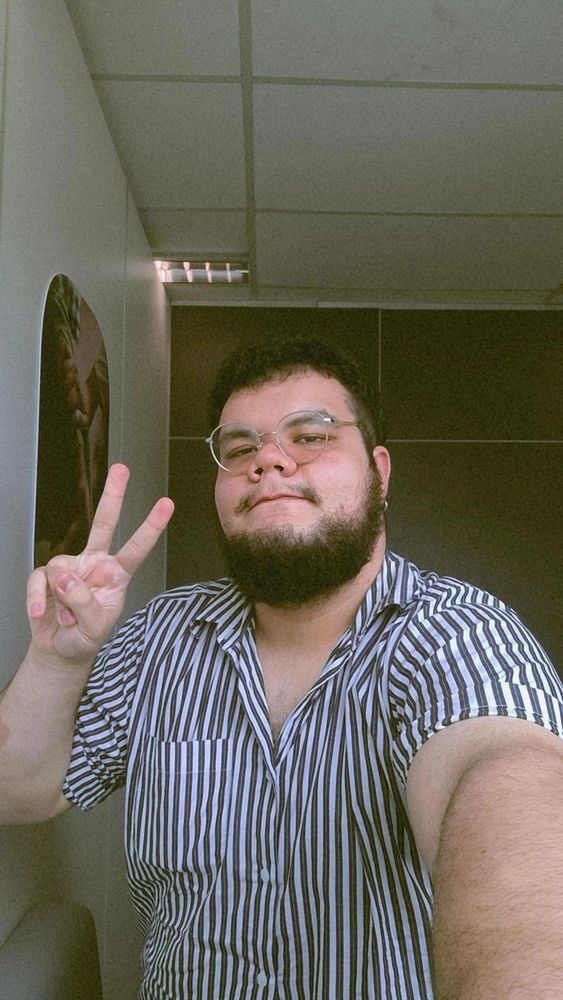 selfie minha (homem branco, gordo, de óculos redondo, barba e cabelos pretos) usando uma camisa listrada preto e branco, segurando o celular com a mão esquerda e fazendo o sinal de paz e amor com a mão direita
