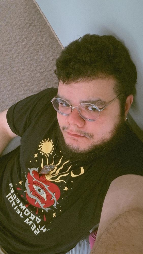 selfie minha (homem branco, gordo, de óculos redondo, barba e cabelo pretos) sentado num sofá, a foto está tirada de cima, mostrando até o começo do meu quadril. estou usando uma camiseta preta com a estampa de um coração vermelho, com um olho no meio, rodeado por espinhos e uma coroa amarela, e embaixo dele está escrito "ah, promessa sem jeito"