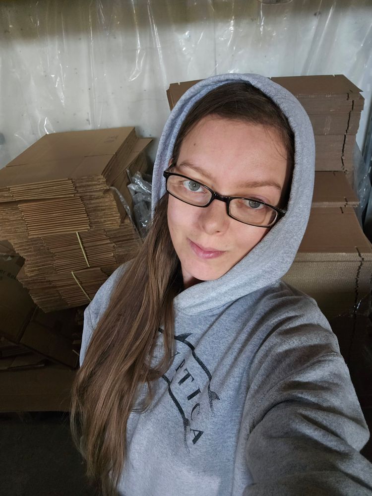 A selfie taken in front of a flat of unfolded boxes. The young woman has rectangular glasses, very long brown hair and a gray Apocalyptica hoodie in with the hood up.