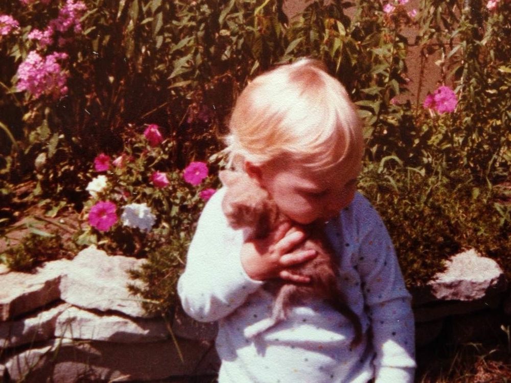 A very blonde, chubby-cheeked toddler is hugging a tiny orange tabby kitten to her cheek. She’s sitting in front of a flower bed with bright pink & white petunias.