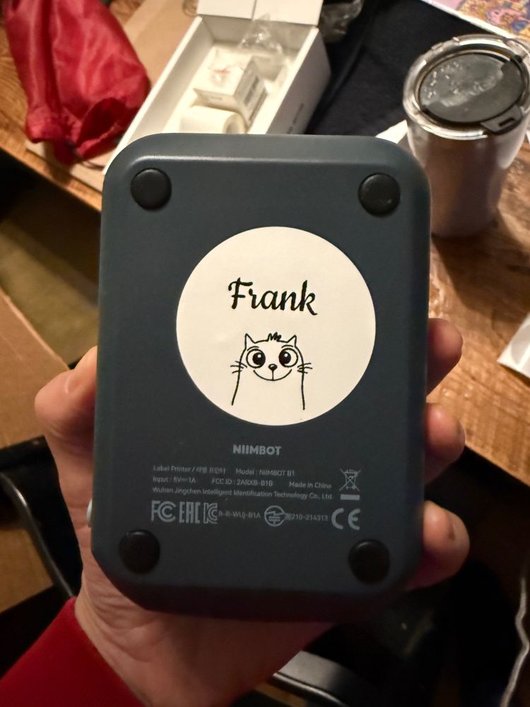 The underside of a small wireless blue label maker, with a round white sticker on it. It says “Frank” and has an icon of a derpy cat.