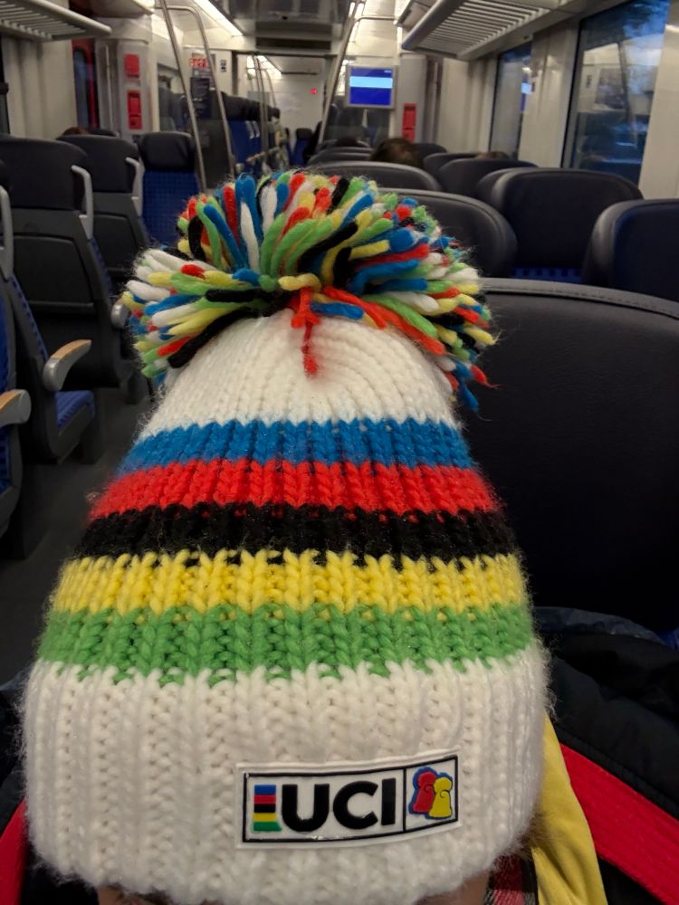 Gestrickte Mütze mit UCI-Regenbogenstreifen und -logo liegt auf einem Sitz im Zug. Dunkelblaue Sitze prägen das Zuginnere – ein Hinweis auf die Reise eines Radsportfans oder Athleten.