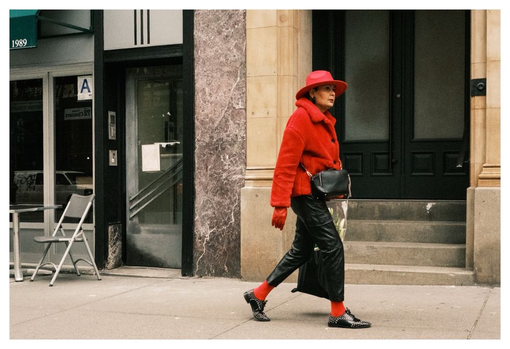 A woman wearing a matching outfit of coat, gloves, socks and a wide brimmed hat in red paired with black leather pants and studded wingtip shoes walks alone on a street in Manhattan. The background contrasts with her in its subdued beige of concrete and marble.