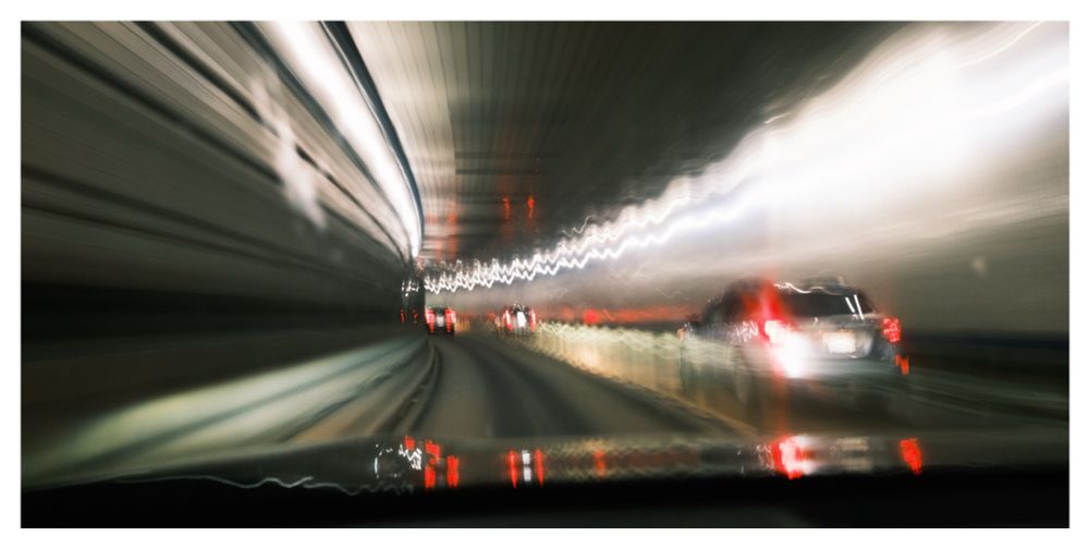 Slow shutter shot through the windshield of a car driving in a tunnel. Streaking lights create an abstract feel as the tunnel curves leftwards in the distance.