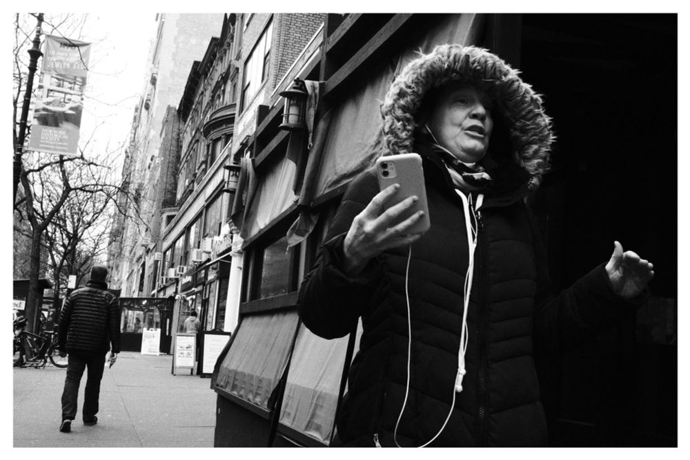 Black and white photo of a New York City street. In the foreground frame right A women in a winter coat is having a serious conversation on the phone, holding her hands up and gesturing. The sidewalk carries into the background behind her in the left side of the frame, a man walks away in the opposite direction.