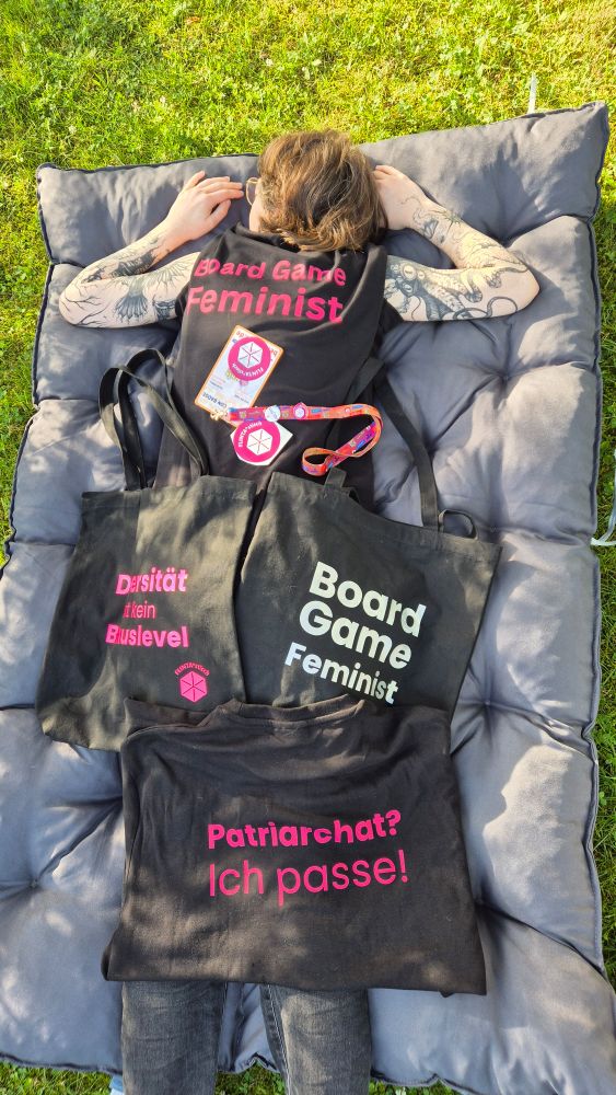 A tattoed woman lying on a grey cushion on the ground. She is wearing a black shirt with pink text that reads "Board Game Feminist". She is covered in black merch (bags, shirts, stickers). Further text on the merch reads "Patriarchat? Ich passe!" and "Diversität ist kein Bonuslevel"
