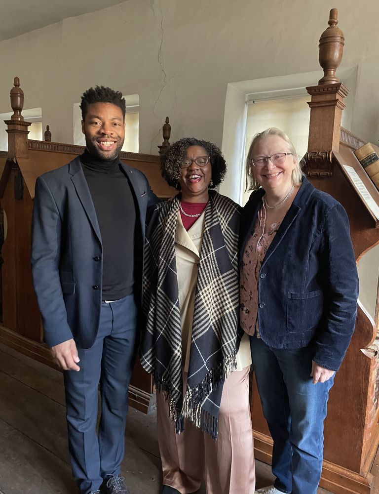 Three people stand together in a warmly lit historic room. The man on the left, wearing a dark suit and black turtleneck, stands with a relaxed posture. The person in the middle, wearing a plaid shawl and a beige outfit, has a bright smile. The person on the right, dressed in a navy blazer over a floral blouse, also smiles warmly. The background features wooden reading stalls with carved finials