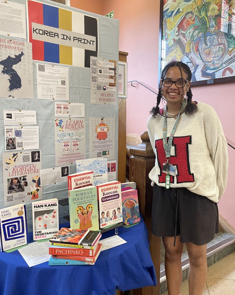 A smiling young woman stand beside a book display containing a selection of Korean novels and language guides