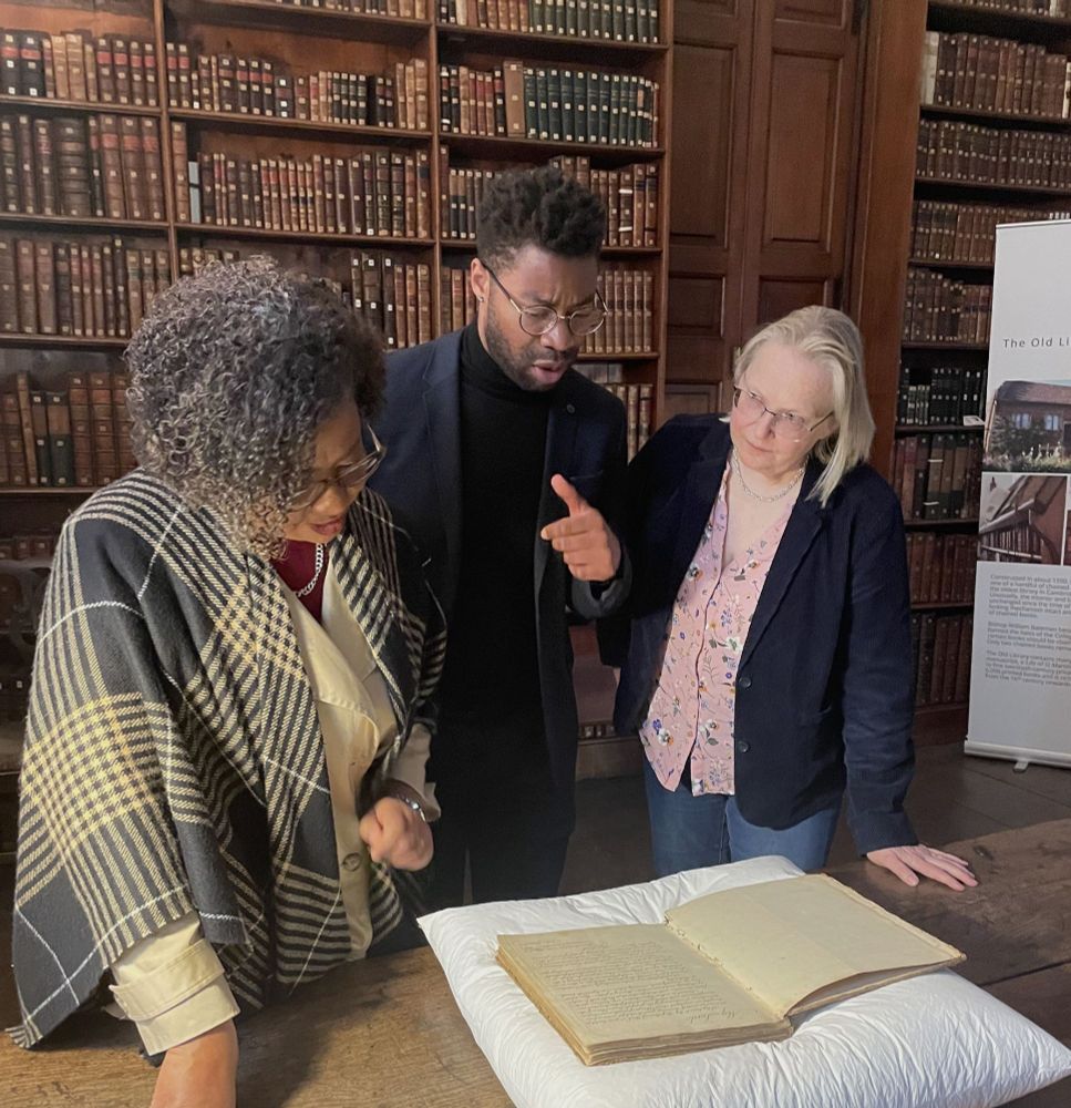 Three people stand around an old manuscript in a historic library setting with wooden bookshelves filled with leather-bound books in the background. The person on the left, wearing a black and white plaid shawl, leans in to examine the book. The person in the center, wearing glasses and a black outfit, gestures animatedly while speaking. The person on the right, wearing a navy blazer and floral blouse, listens intently with her hand resting on the table. The book is placed on a white cushion, suggesting careful handling of the historical document.
