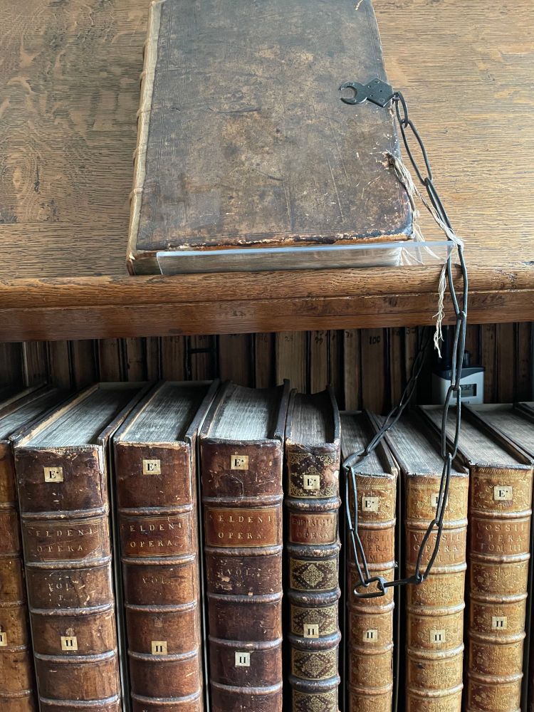 Shelf of books in Trinity Hall's Old Library. The books are in brown leather bindings beneath a shelf containing one chained book