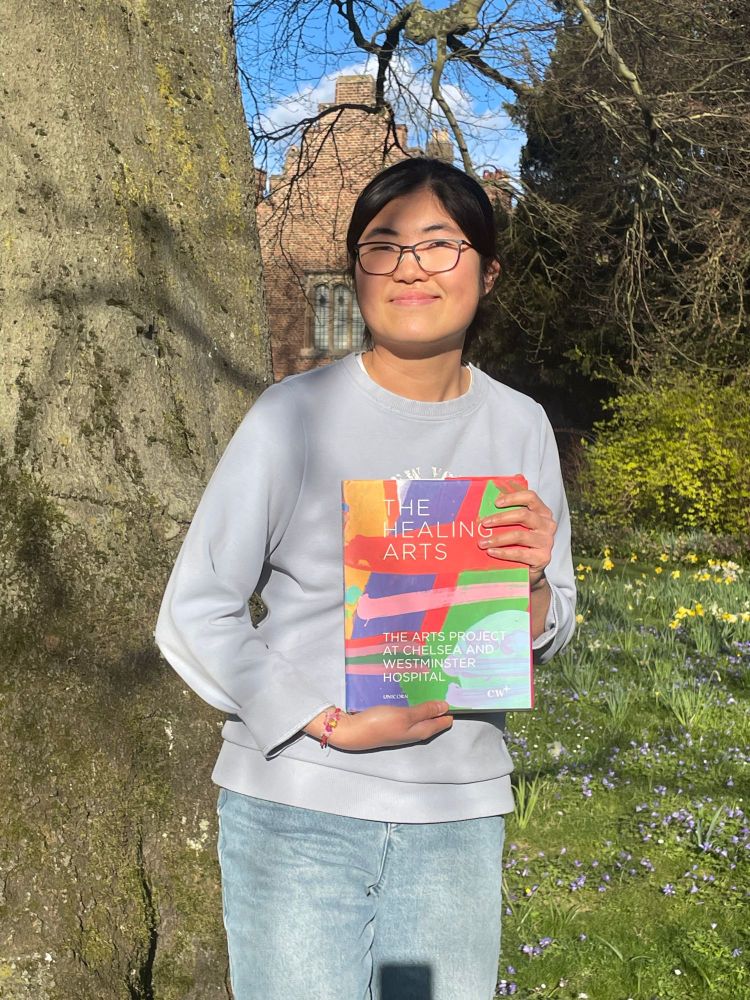 A young woman with glasses and dark hair tied back stands outdoors in sunlight, smiling gently. She is wearing a light gray sweatshirt and blue jeans, holding a colorful book titled The Healing Arts: The Arts Project at Chelsea and Westminster Hospital. 