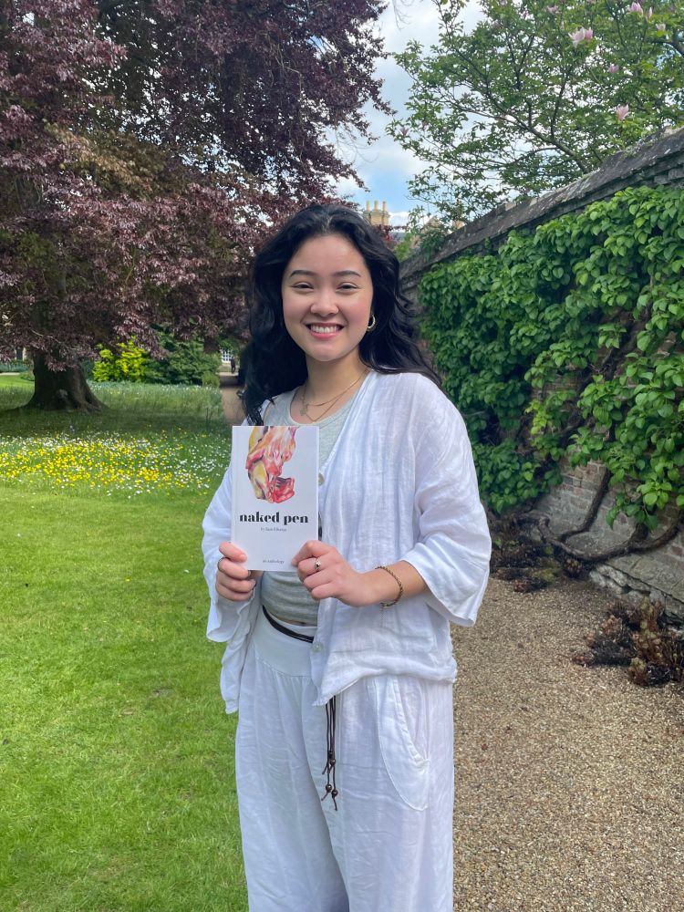 A young woman with dark hair is smiling while holding a book entitled "Naked pen". She is in a garden with a tall tree in the background