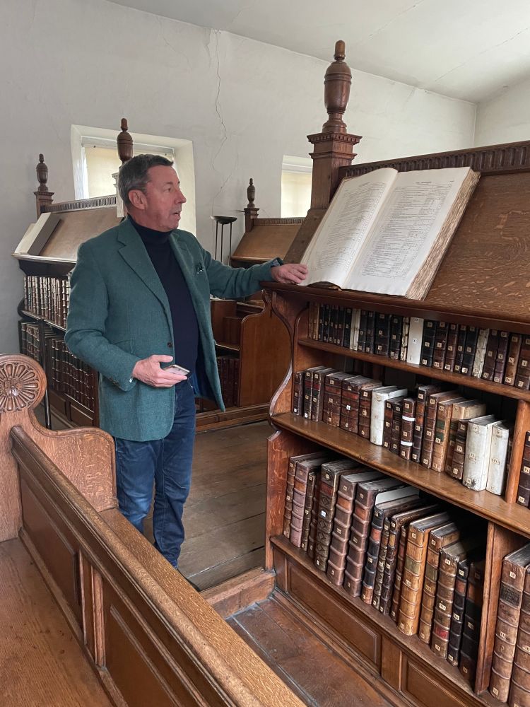 A man stands next to a wooden lectern on which he looks at an open book on the sloping top shelf. More books are on the shelves below.